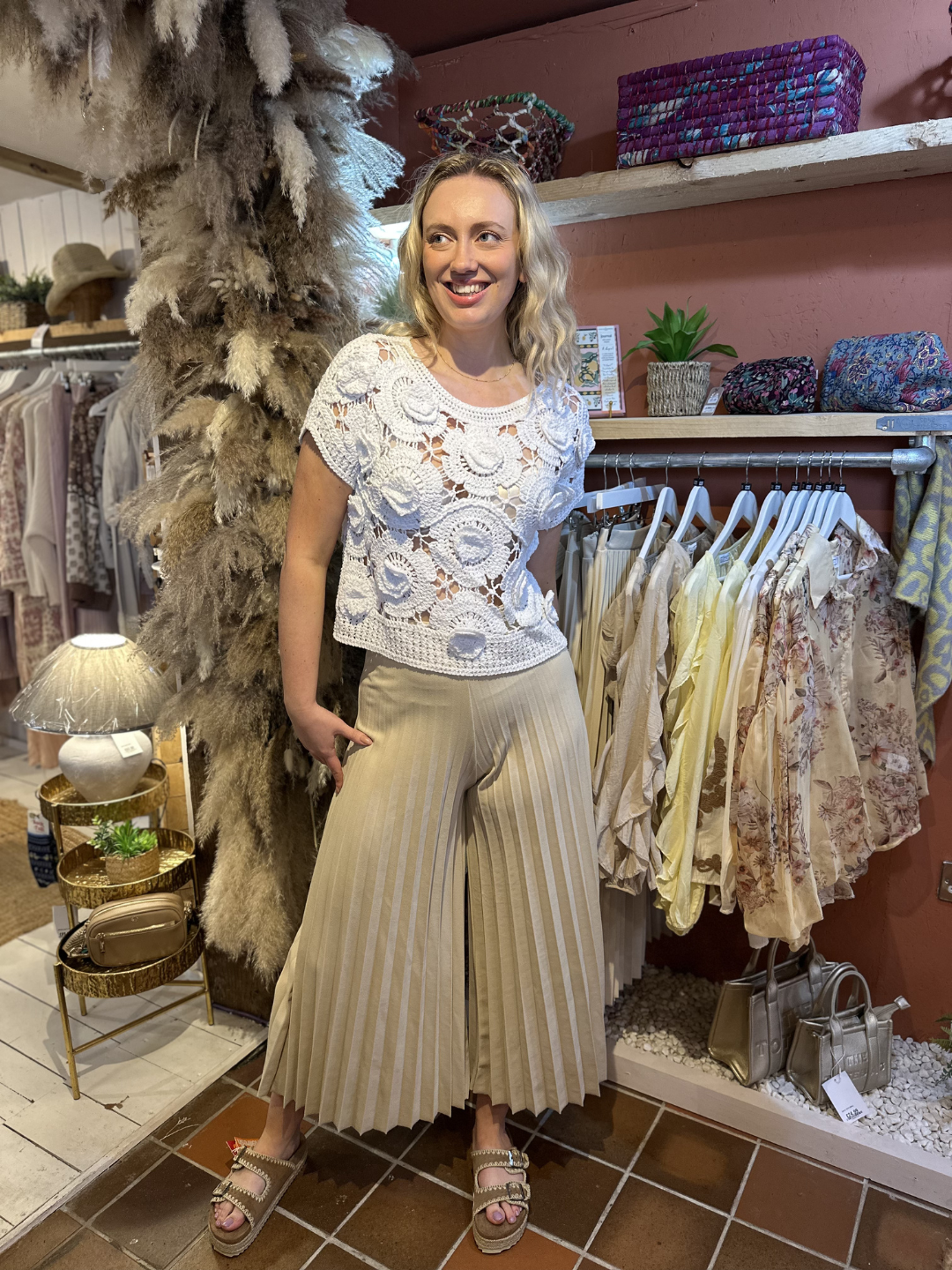 A woman stands in a boutique, wearing a sheer knitted jumper as a spring layering piece, beige pleated culottes and sandals. She is smiling, surrounded by clothing rails and boutique décor.