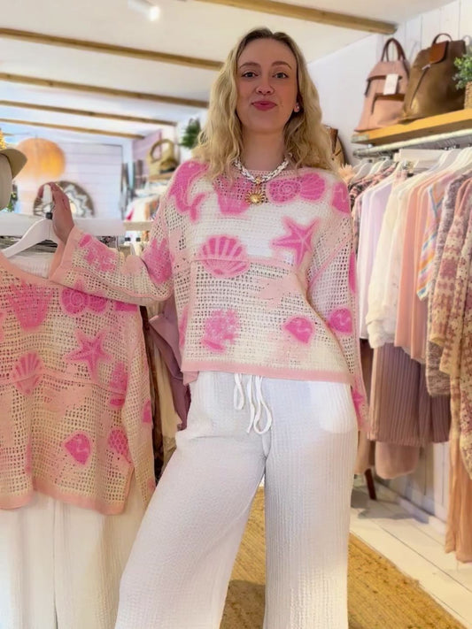 A woman stands in a clothes shop wearing a white and pink ocean-inspired jumper and white trousers, holding a matching jumper on a hanger. Clothing rails and accessories are visible in the background.