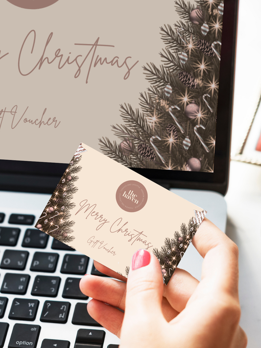 A hand holds a Merry Christmas Haven Fashion & Lifestyle e-gift card featuring a festive pine and bauble design, in front of a laptop showing a matching Christmas-themed graphic.