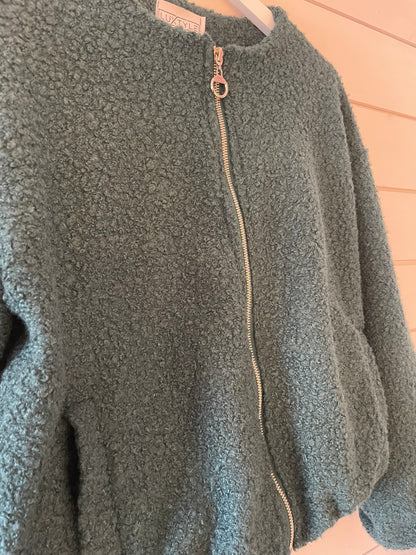 A textured, light green sustainable fashion zip-up jacket hangs on a hanger against a light wooden wall.