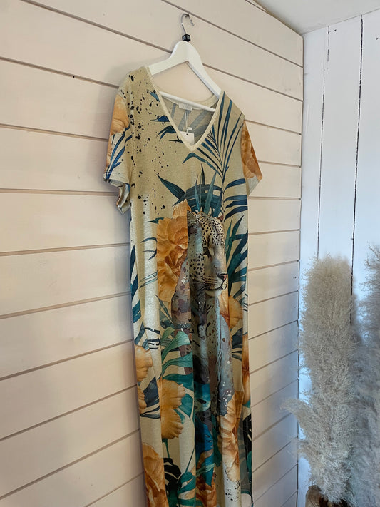 A short-sleeved, V-neck evening dress with a jungle print, displaying large yellow flowers, green leaves, and a leopard, hangs on a white hanger against a wooden-panelled wall.