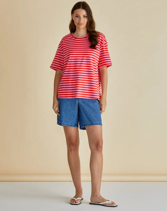 A woman stands facing forward, dressed in a boxy T-shirt with red and white stripes, denim shorts, and white sandals against a plain beige background.