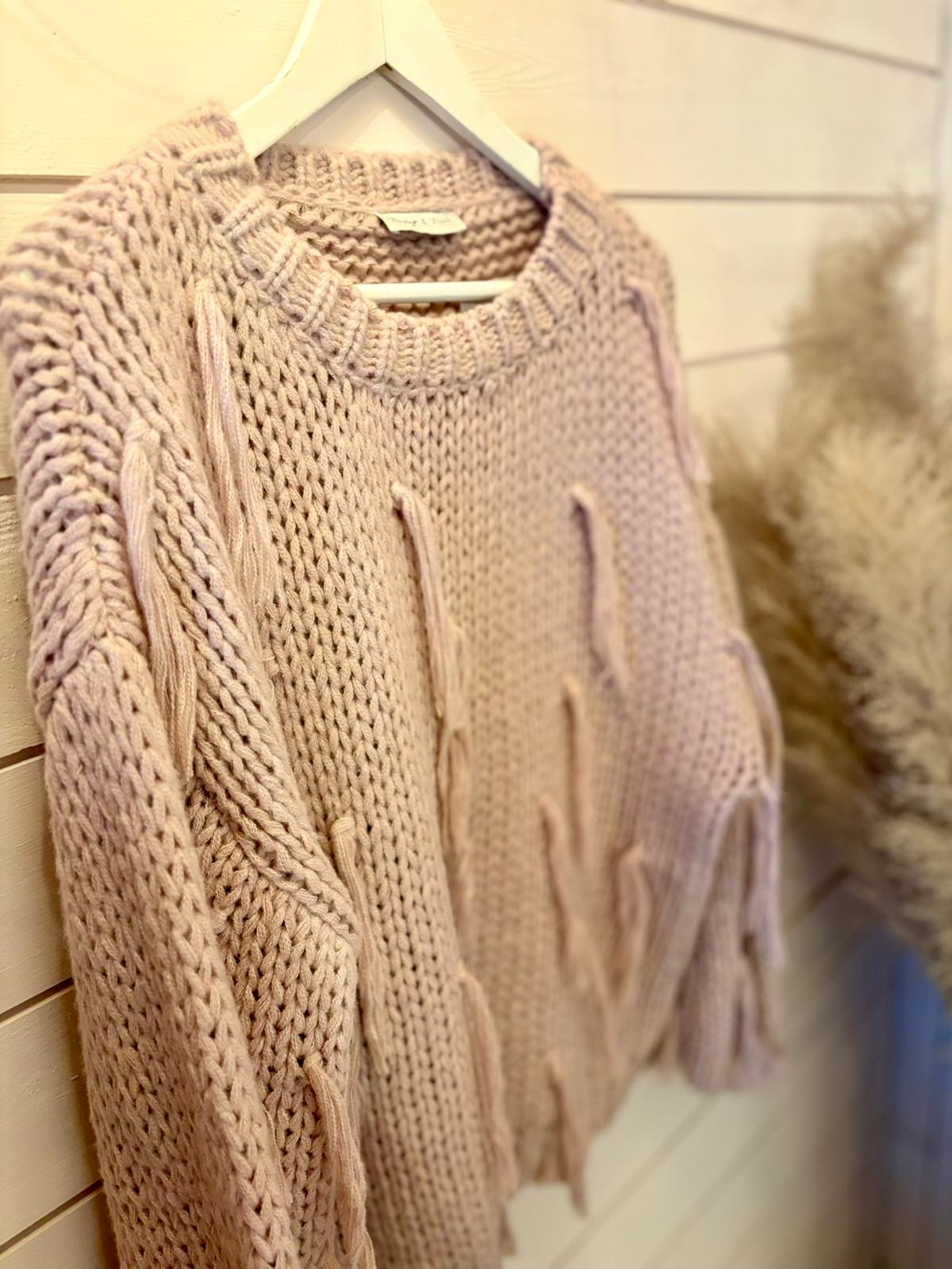 A chunky, light pink jumper with cable details hangs on a white hanger against a wooden wall, ideal as a cosy vegan jumper for chilly days.