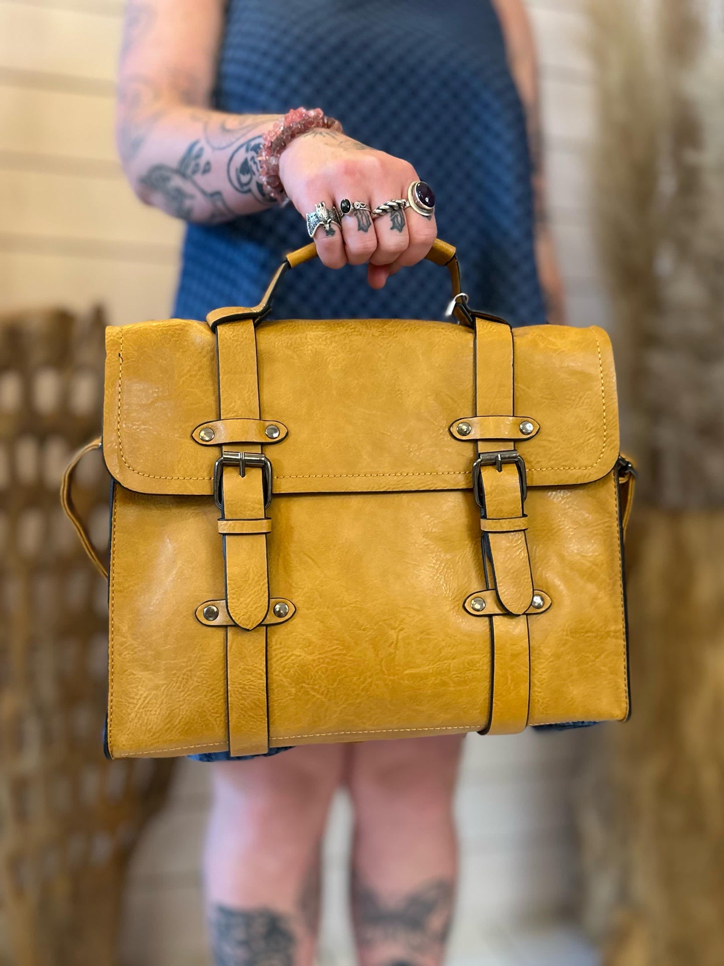 A person with tattoos holds a vegan-friendly yellow faux leather satchel by the top handle, featuring two front buckle straps that elevate their business look.