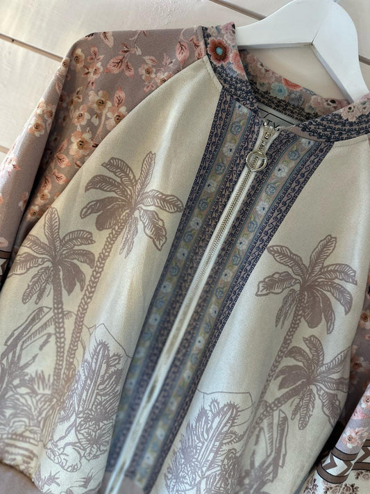 A close-up of a light-coloured jacket with a zip, featuring palm tree prints and floral-patterned sleeves, hanging on a white hanger.