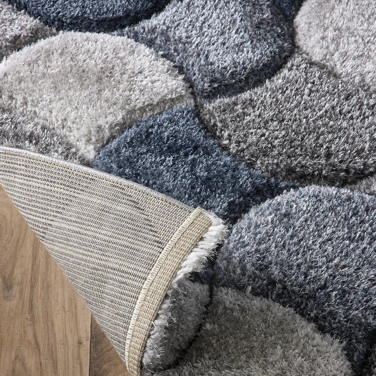 Close-up of a corner of a contemporary rug in various shades of grey and blue on a wooden floor, with the corner folded over to reveal the hardwearing polyester rug backing.