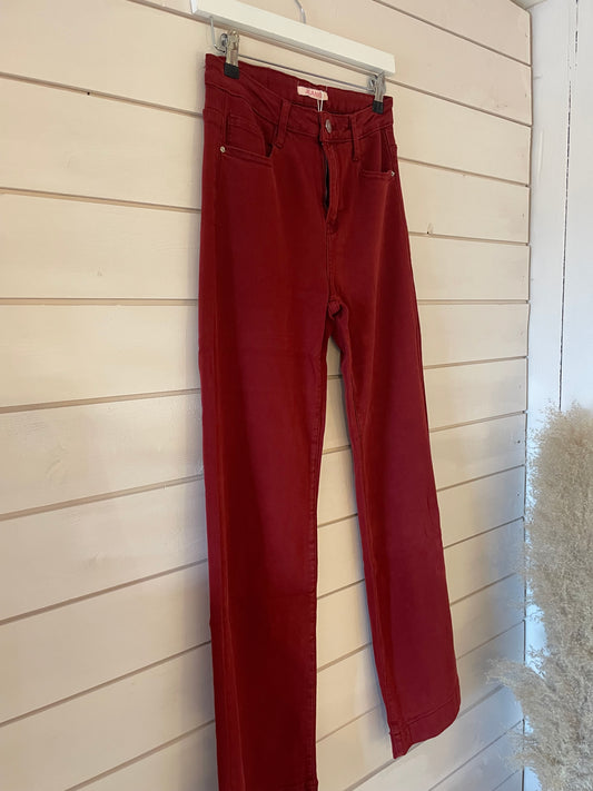A pair of wide-leg red jeans hangs on a clear hanger against a light wood-panelled wall, making a bold statement in fashion-forward denim.