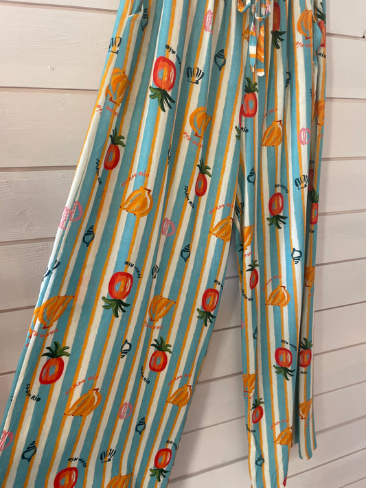 Lightweight trousers featuring vertical blue and white stripes, cheerful fruit motifs, and abstract designs—made from vegan-friendly fabric for easy casual wear. Displayed stylishly against a white wooden wall.