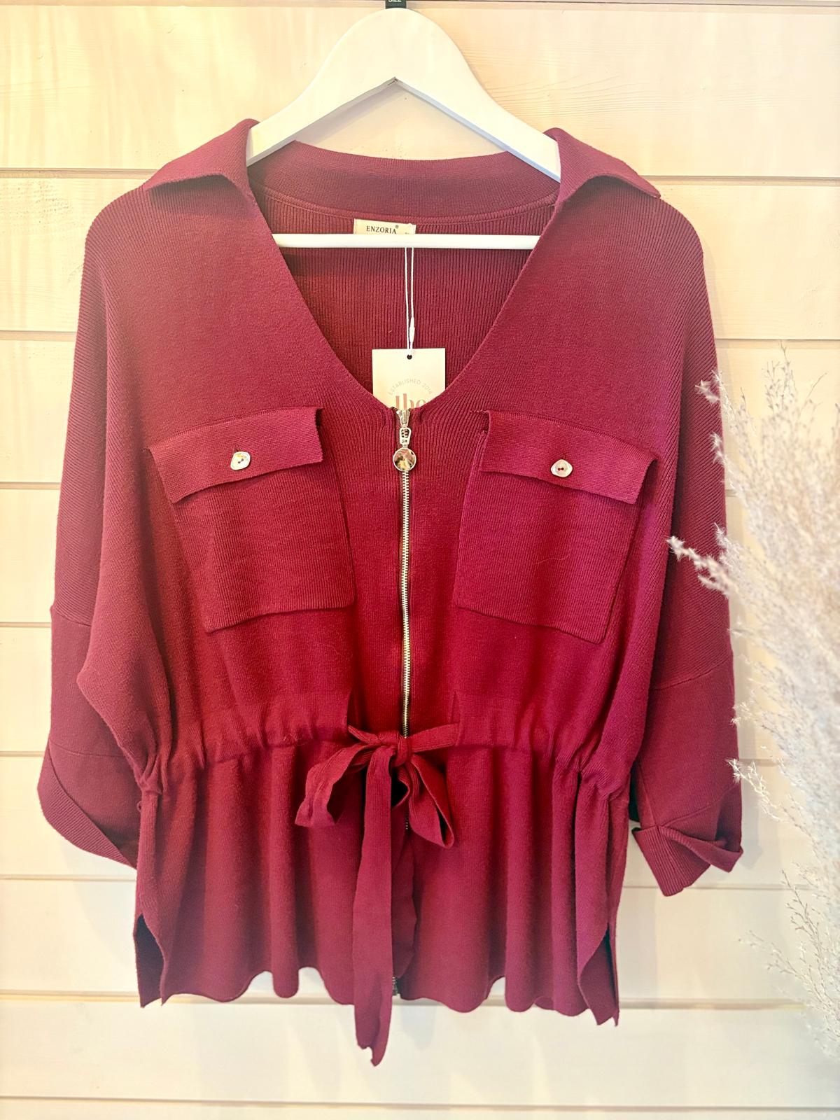 A maroon, long-sleeved blouse with a zip front, two flap chest pockets, a collar, and a waist tie, displayed on a white hanger against a light wooden background.