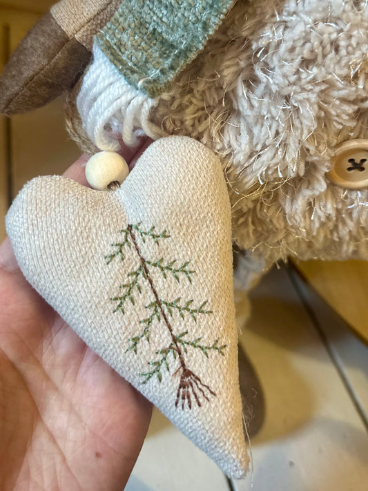 A hand holds a fabric heart ornament featuring a green embroidered branch, beside a textured soft toy with a button detail—ideal festive décor for creating a cosy atmosphere indoors or outdoors at Christmas.