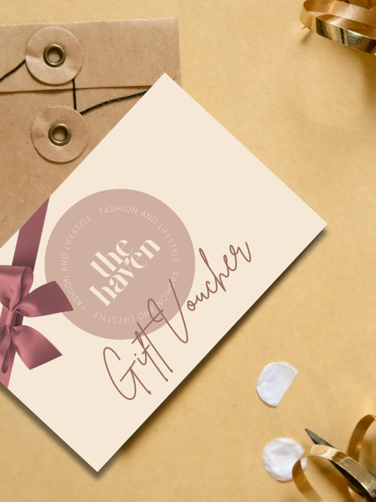 A Haven Fashion & Lifestyle gift card lies on a beige surface, with an envelope, ribbon, white petals, and scissors nearby—ideal for unforgettable fashion presents.