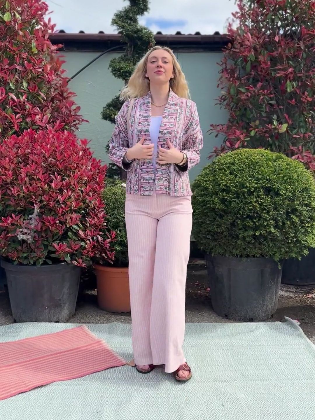 A woman stands outside on a rug, dressed in pink pinstripe wide-leg jeans, a patterned pink jacket, and sandals, with green and red foliage in large pots behind her.