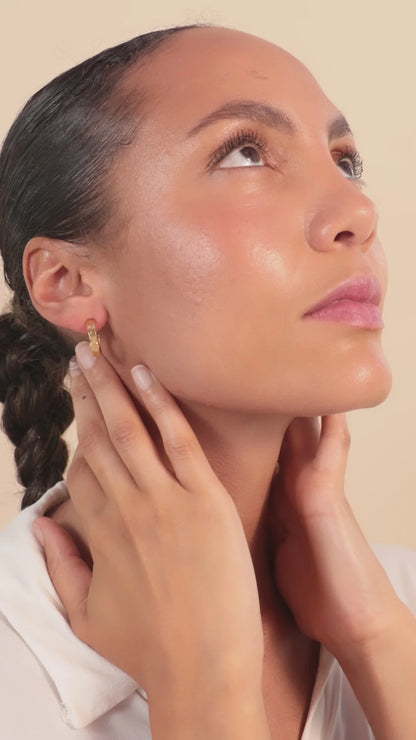 Woman with plaited hair looks upwards, touching her neck with both hands. She is wearing a white top and gold crescent hoop earrings, set against a beige background.