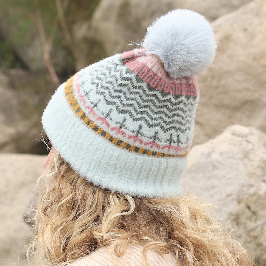 Person with curly blonde hair wearing a cruelty-free, multi-patterned knitted hat featuring a pom-pom and geometric designs in white, pink, mustard, and grey, outdoors near large rocks.