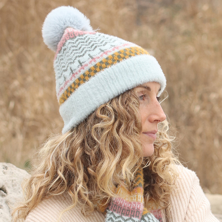 Woman with curly blonde hair wearing a vegan hat with a multi-patterned design and pom-pom, along with a matching scarf, sitting outdoors with a neutral expression.