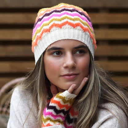 A woman wears a vegan-friendly chevron beanie hat and matching zigzag fingerless gloves, with a neutral backdrop of wooden planks.