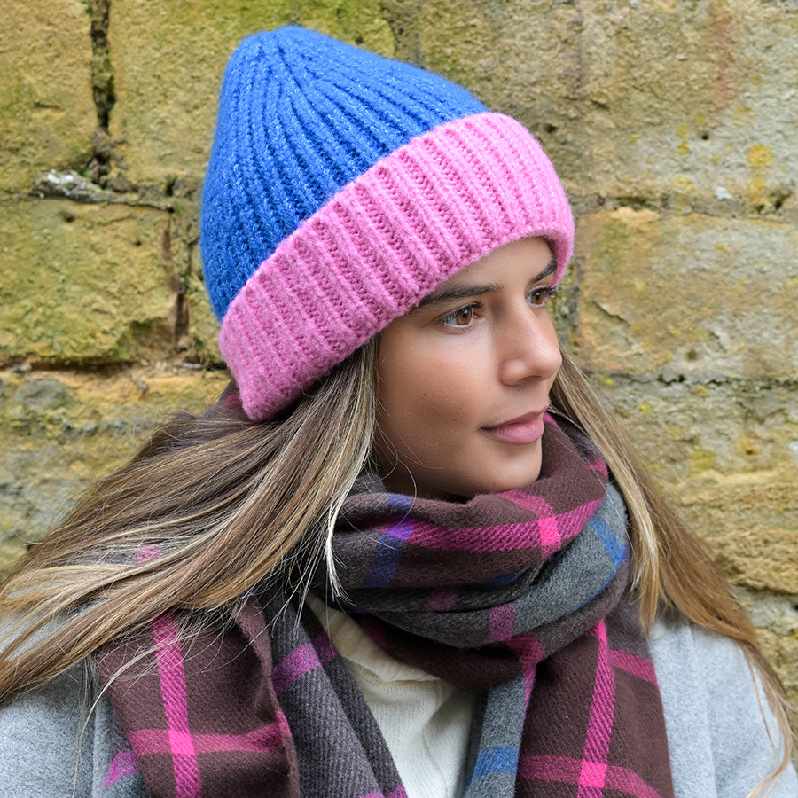 A person in a blue and pink vegan knitted beanie and a brown checked scarf stands before a stone wall, gazing to the side. This cosy winter essential brings warmth and style to any chilly day.