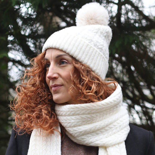 A woman with curly red hair wearing a cream knitted beanie made from recycled vegan yarn and a matching scarf, standing outdoors with trees in the background.