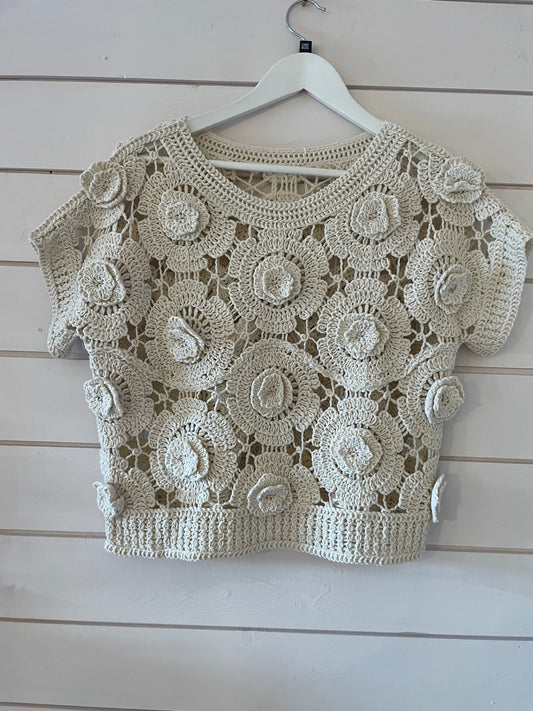A cream floral jumper with short sleeves and an intricate crochet pattern, ideal as a sheer layering piece for spring fashion, shown hanging on a hanger against a white painted wooden wall.