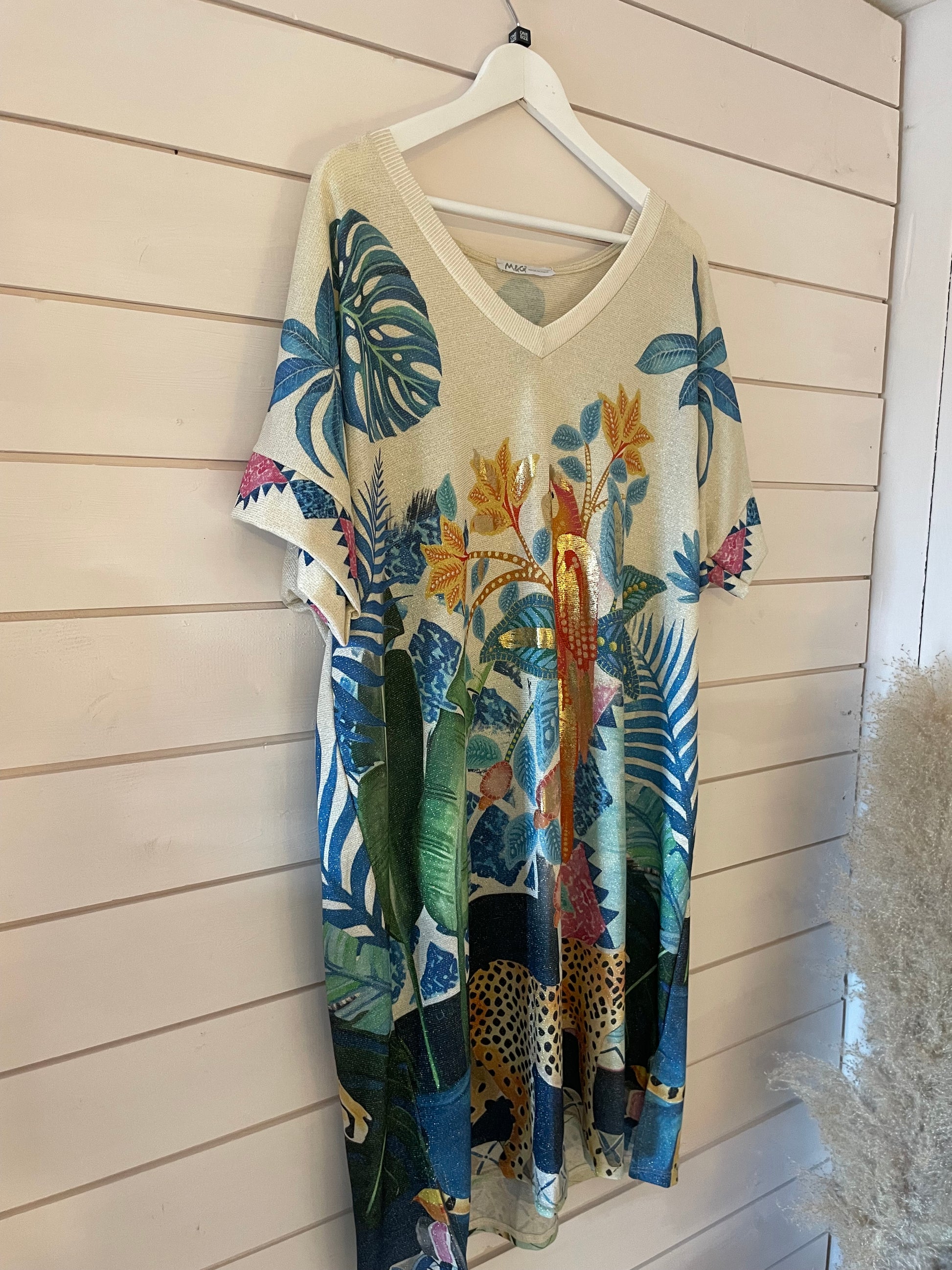 A short-sleeved, vegan-friendly dress with a V-neck hangs on a white hanger; it features a colourful tropical print with leaves, flowers and leopards.