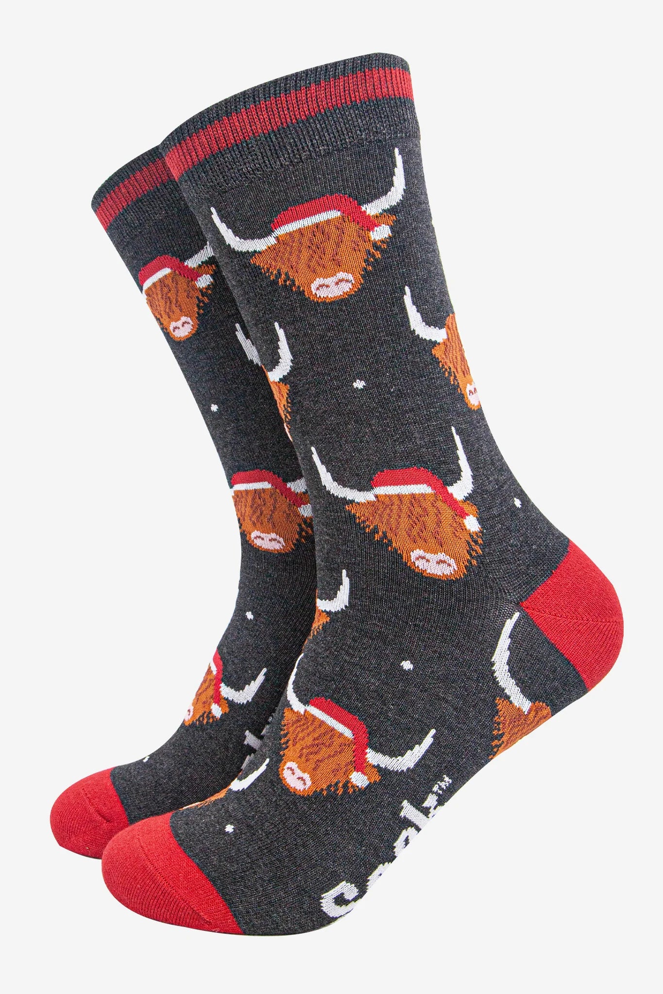 A quirky pair of dark grey socks with red heels, toes, and cuffs, adorned with cartoon cows in Santa hats and small white spots like snow. These vegan-friendly socks are ideal for adding festive cheer!.
