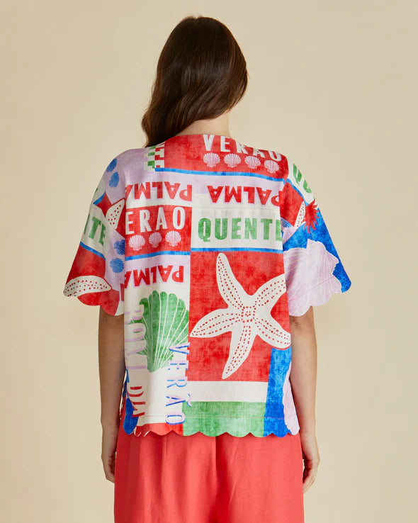 A person with long brown hair, viewed from behind, wears a colourful seashell-print shirt with scalloped edges over red shorts—showcasing bold graphics and sustainable fashion against a beige backdrop.