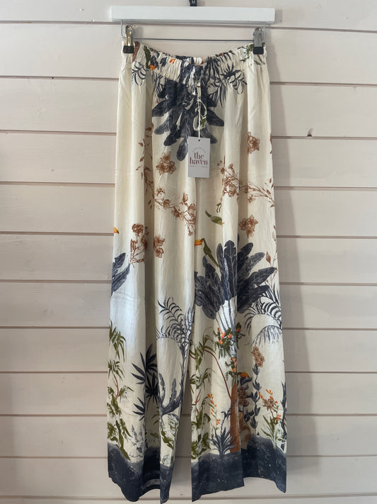 Wide-leg cream trousers in lightweight fabric, featuring a botanical and bird print, with elasticated waistband and drawstring, hang on a white hanger against a light wooden wall. Ideal as stylish Blue jungle trousers.