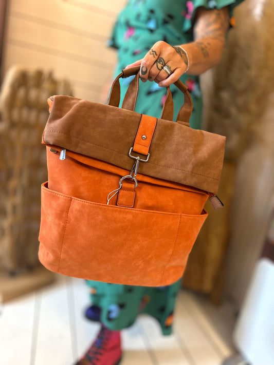 A person with tattoos holds a luxury rucksack made of orange and brown suede with front pockets and a clasp fastening, showcasing sustainable fashion while standing indoors.