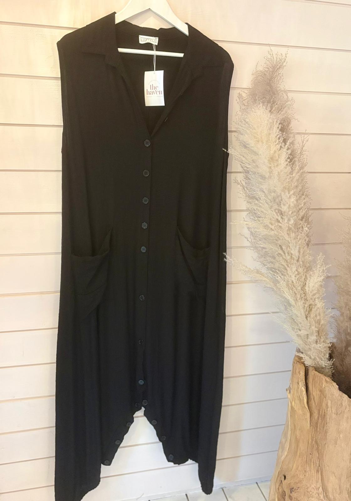 A sleeveless black button-up jumpsuit dress with large front pockets hangs on a wooden hanger against a light wood-panelled wall; decorative pampas grass can be seen on the right.