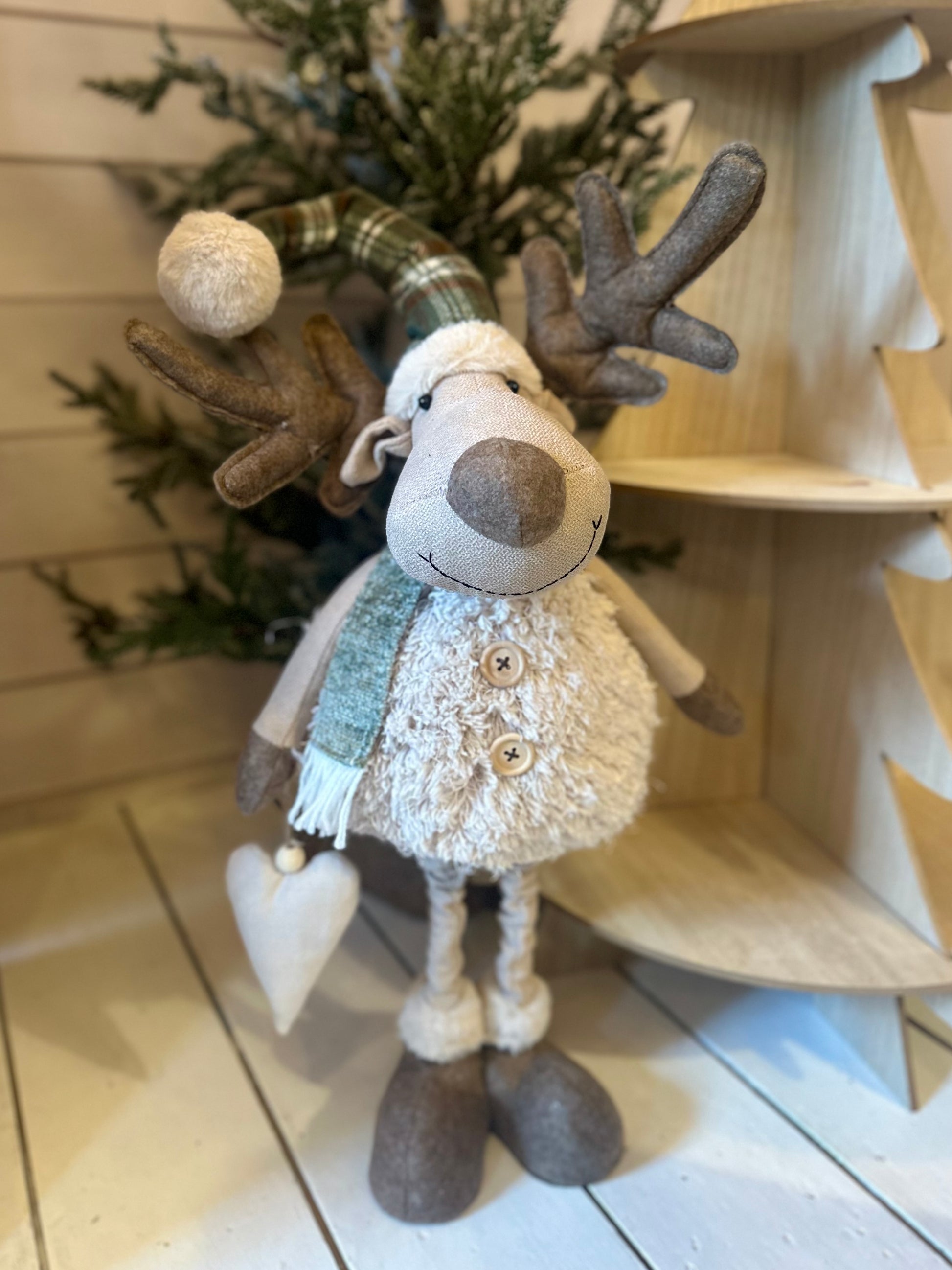 Plush reindeer figurine wearing a scarf and hat, holding a heart ornament, stands before a small artificial tree and wooden decorations—ideal festive décor for your home.