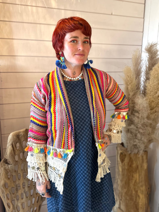 A person with short red hair is wearing a blue checked dress and a bohemian, colourful striped cardigan with tassels and lace, standing indoors near dried plants.