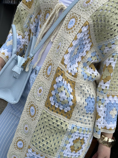 Person wearing a crocheted patchwork cardigan in pastel shades, a light blue crossbody bag, white shirt, blue and white striped trousers, and a gold watch.
