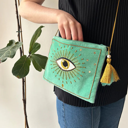 A person in blue jeans and a black top holds the Glimmer Crossbody Bag—a vegan-friendly accessory featuring an embroidered eye, gold accents, and a yellow tassel—beside a leafy plant.