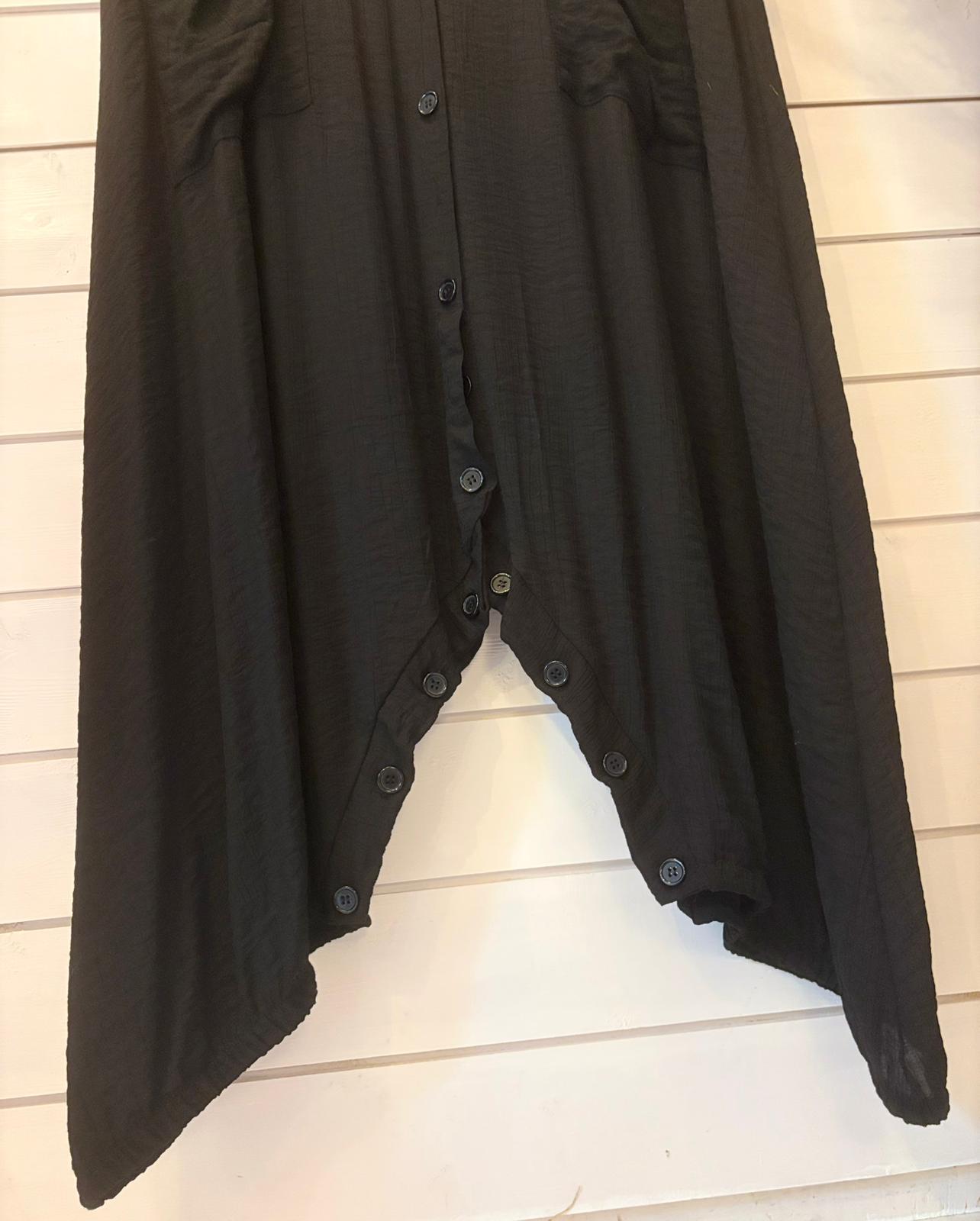 Black jumpsuit dress with an asymmetrical V-shaped hem and central buttons, displayed against a white wooden wall. This versatile jumpsuit provides a stylish, one-size fit for effortless elegance.