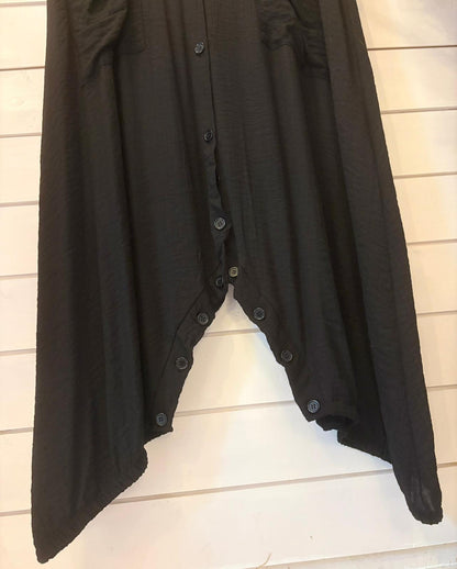Black jumpsuit dress with an asymmetrical V-shaped hem and central buttons, displayed against a white wooden wall. This versatile jumpsuit provides a stylish, one-size fit for effortless elegance.