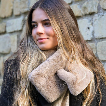 A woman with long, wavy hair wears a black coat and a taupe faux fur pull-through scarf, standing before a stone wall—a luxurious winter accessory.