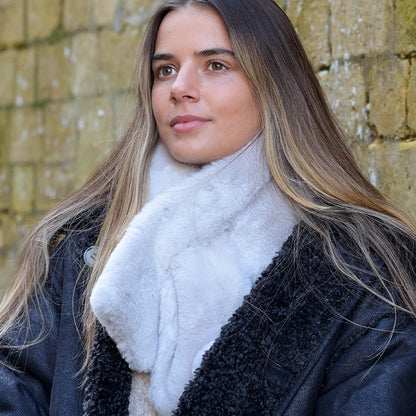 A woman with long hair, dressed in a black coat and a luxurious white pull-through scarf, stands in front of a stone wall.