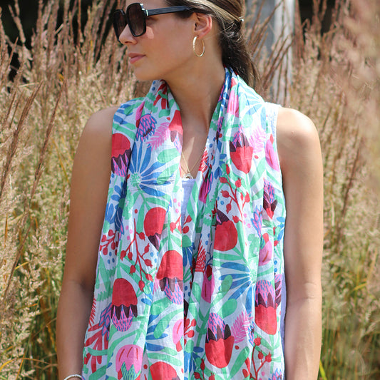 Wearing sunglasses and a vibrant meadow print scarf, the person stands outdoors among tall grass, looking to the side.