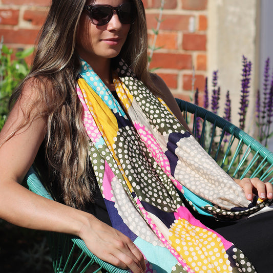 A woman with long hair and sunglasses sits outdoors in a turquoise chair, her colourful Bamboo Scarf with a spotty circle print adding a vibrant touch against the brick wall backdrop.