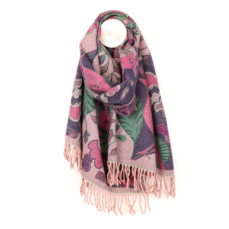 A luxurious jacquard scarf with a floral pattern and fringed ends, featuring a reversible pink mix, hangs draped over a white peg.