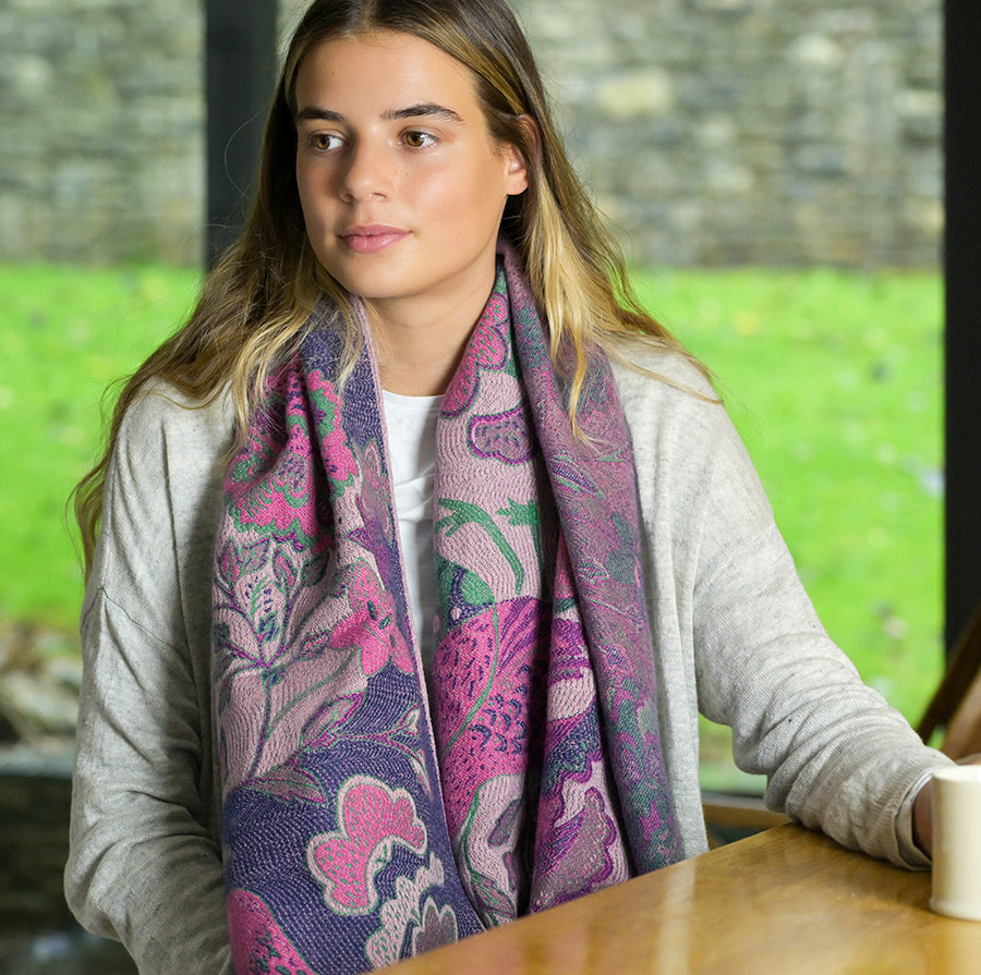 A woman sits inside, wrapped in a jacquard scarf with luxurious fringed ends. Her colourful patterned scarf complements her light grey jumper, while a cup rests on the wooden table in front of her.