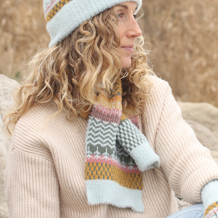 A woman with curly blonde hair wears a light blue knitted hat and matching vegan scarf with a colourful multi-patterned design, paired with a beige jumper, sitting outdoors near rocks.