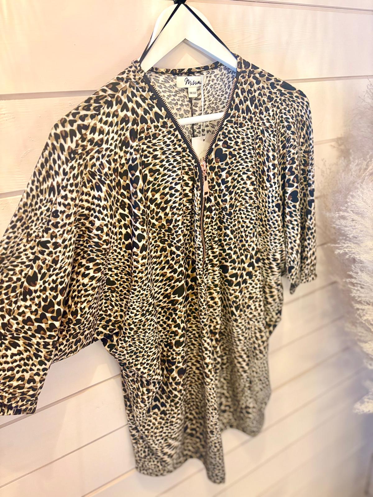 A leopard print blouse with a front zip is hanging on a white hanger against a white wooden wall.