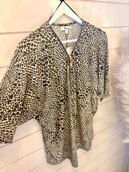 A leopard print blouse with a front zip is hanging on a white hanger against a white wooden wall.