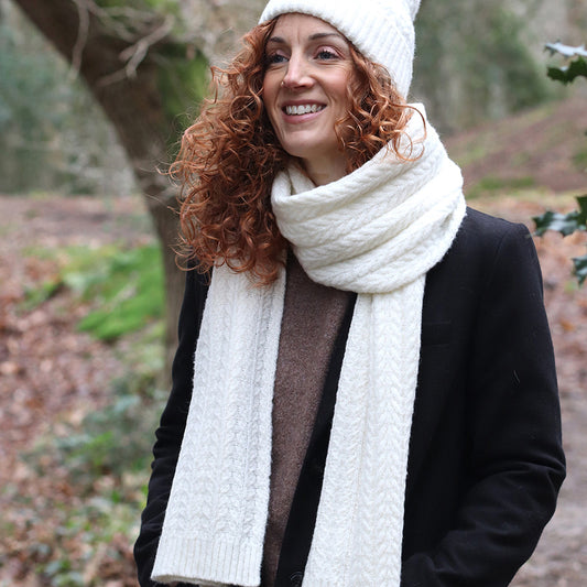 A woman in a cosy knitted scarf and hat with a black coat stands outside in a wooded area, smiling.