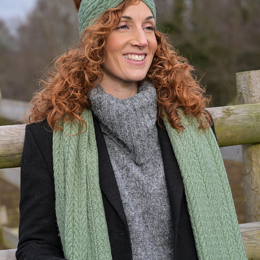 A woman with curly red hair in a green knitted headband and a sage scarf, made from recycled vegan yarn, stands outdoors by a wooden fence wearing a grey turtleneck jumper and black coat.