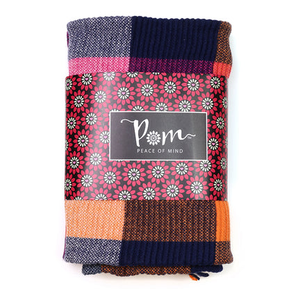 A folded multicoloured knitted blanket with a floral-patterned label bearing the words POM Peace of Mind, ideal as a stylish accessory or a cosy alternative to a rust and pink scarf.