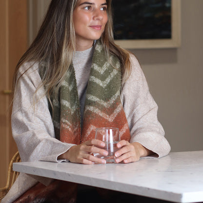 A woman wearing a cosy scarf and light jumper sits at a table, holding a clear glass of water with both hands.
