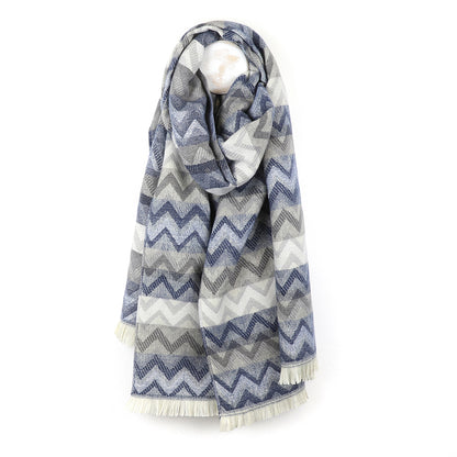 A long chevron scarf featuring a blue, grey, and white zigzag pattern is draped on a white background; the ends are finished with short fringe, making it a vibrant fashion accessory.