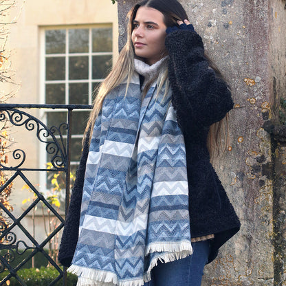 A woman with long hair, dressed in a thick black coat and a vibrant chevron scarf, stands outdoors near a stone wall and iron gate.